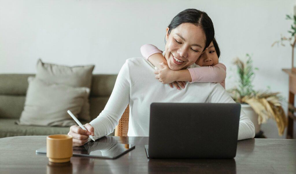 Woman working on laptop with child hugging neck