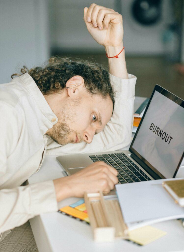 Man with head resting on desk alongside a laptop with a screen that reads "Burnout."
